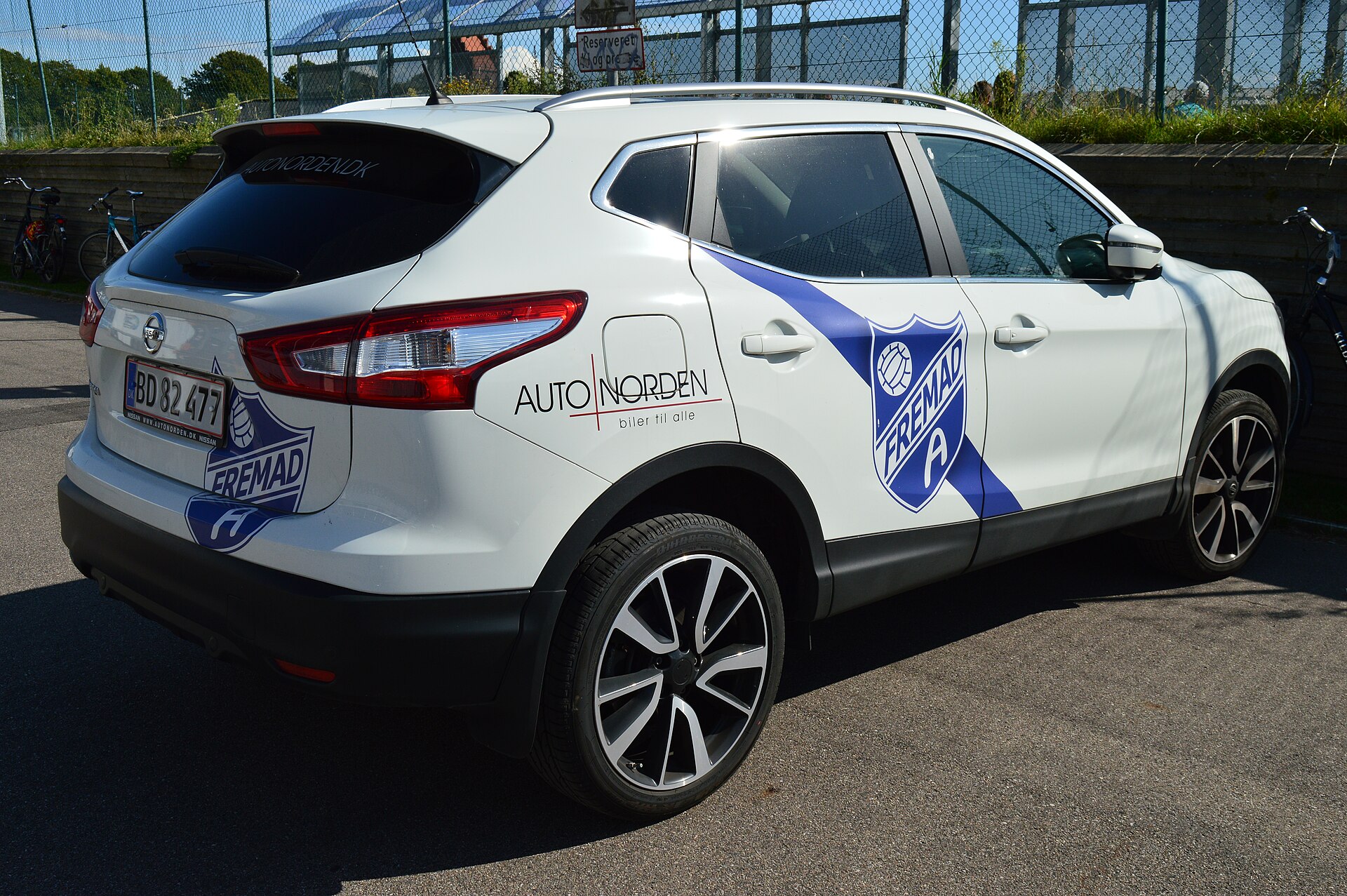 A sponsored car related to the Danish association football club BK Fremad Amager, viewed from the back, parked at Sundby Idrætspark (south of the grand stand), provided by Autonorden. The white coloured (with a blue stripe) Nissan car featured the club's logo on the sides and back witht logo of the 