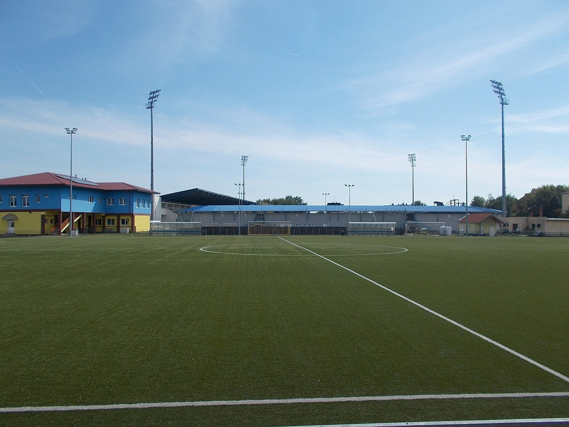 : Sport facility, in front a grassy city stadium. At back behind blue roof is the stadium with grandstands. Viewed from Kavicsos-tó street. - Entry from Széchenyi István street, Mezőkövesd , Borsod-Abaúj-Zemplén County , Hungary .