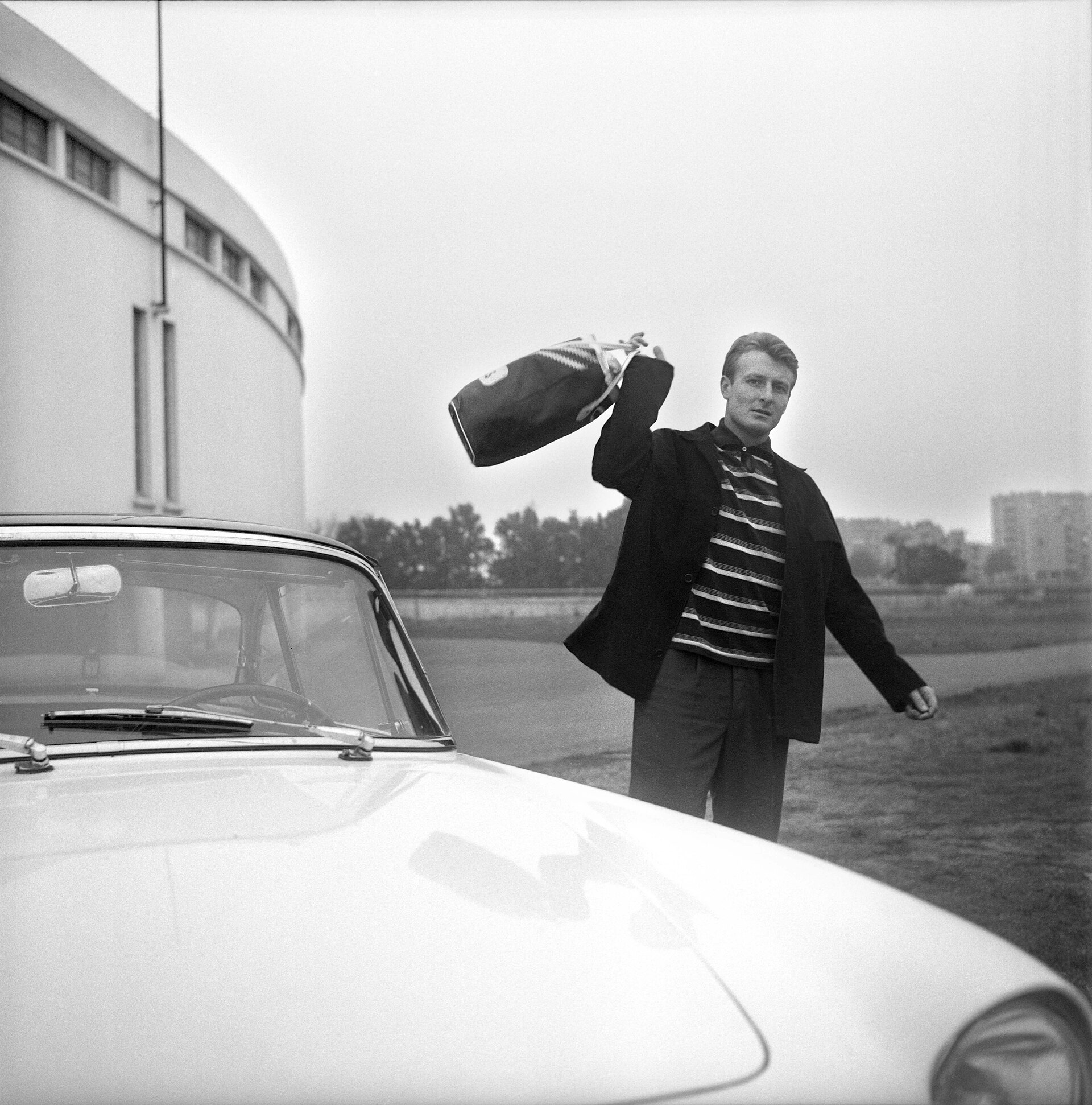 Stadium municipal de Toulouse, 1bis allées Gabriel-Biénès. Entre 15 et 18 octobre 1962. Jacky Bernard (joueur du Toulouse Football Club) à côté de sa voiture, tenant un sac de sport dans une main (vue de face) ; en arrière plan le stade. .