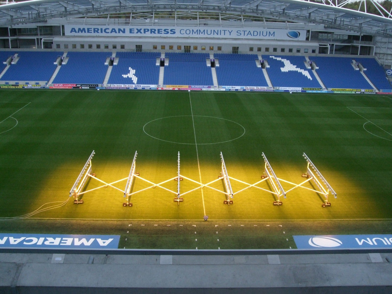 Pitch & East Stand - Amex Stadium. Viewed from West Stand, the lighting on the pitch is part of the club's commitment to environmentl sustainability. – American Express Community Stadium. After many years without a permanent home, Brighton and Hove Albion's new ground at Falmer was opened in August