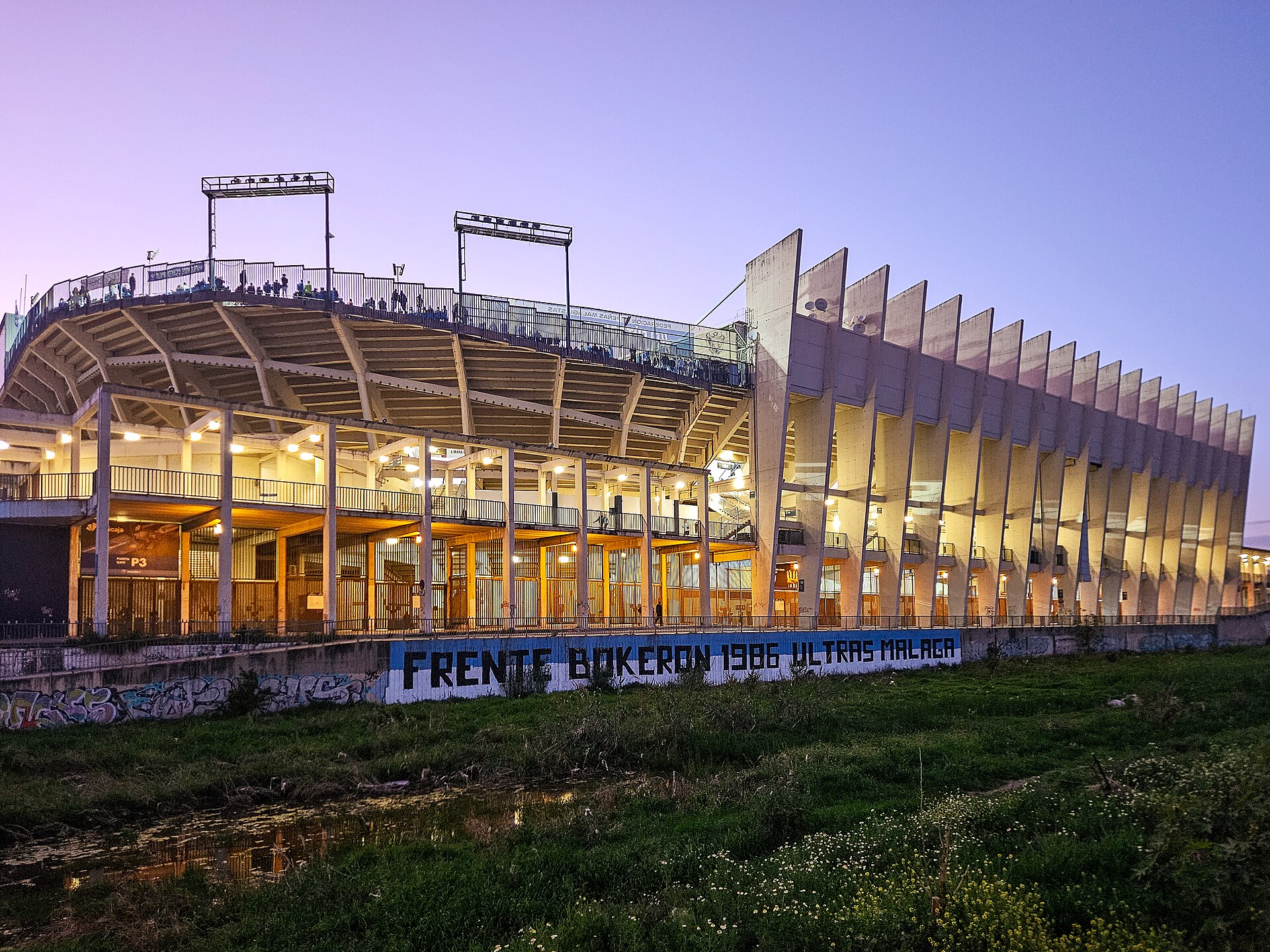 Estadio La Rosaleda, Málaga, as seen from the southeast across the Río Guadalmedina. The stadium lights are on as a game is in progress.