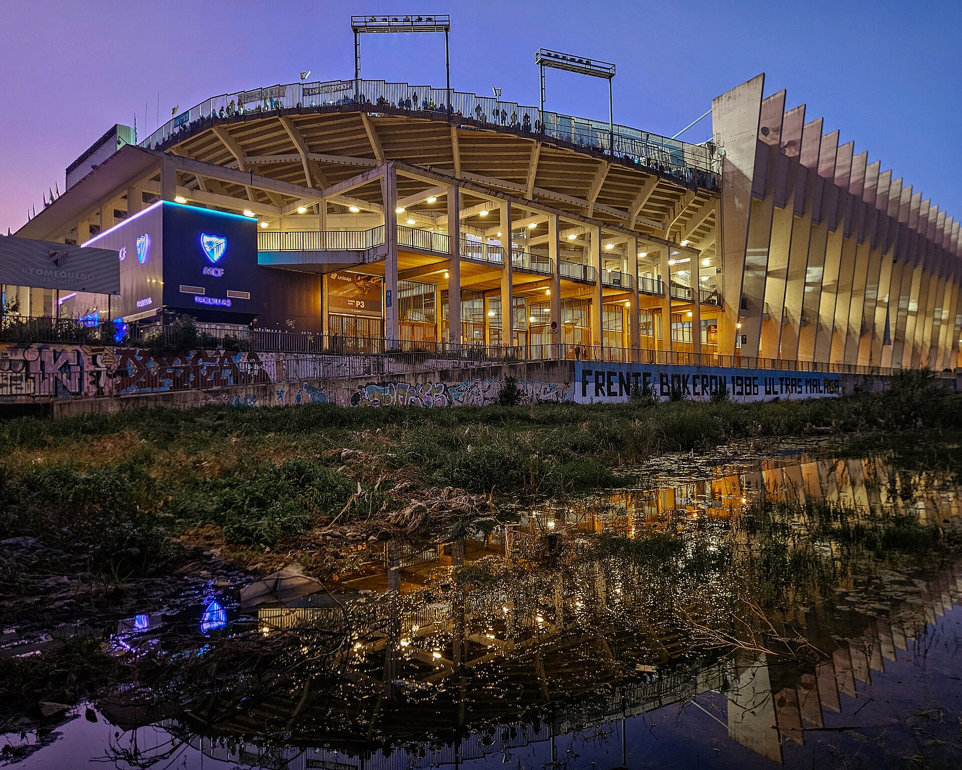 Estadio La Rosaleda, Málaga, from the left bank of the Rio Guadalmedina. The stadium lights are on as a Málaga CF home game is in progress.